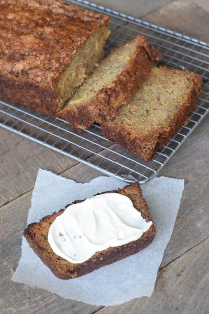 Zucchini Pineapple Bread sliced on a wire rack. One slice toasted and spread with cream cheese on parchment paper