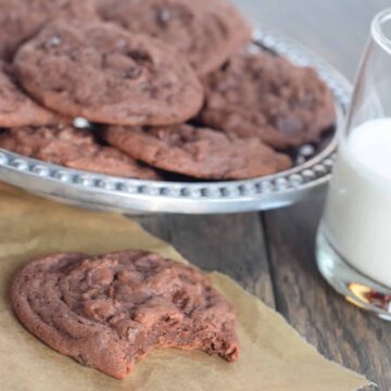 Chocolate cookies with chocolate chips with bite out of one on parchment. Plate of them behind it and glass of milk