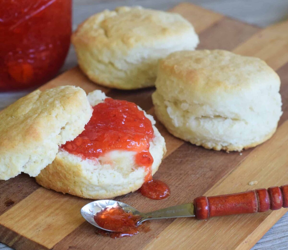 Strawberry Jam spread on 2 Ingredient Biscuits with jam spilling out of a little wooden spoon on a wooden cutting board.