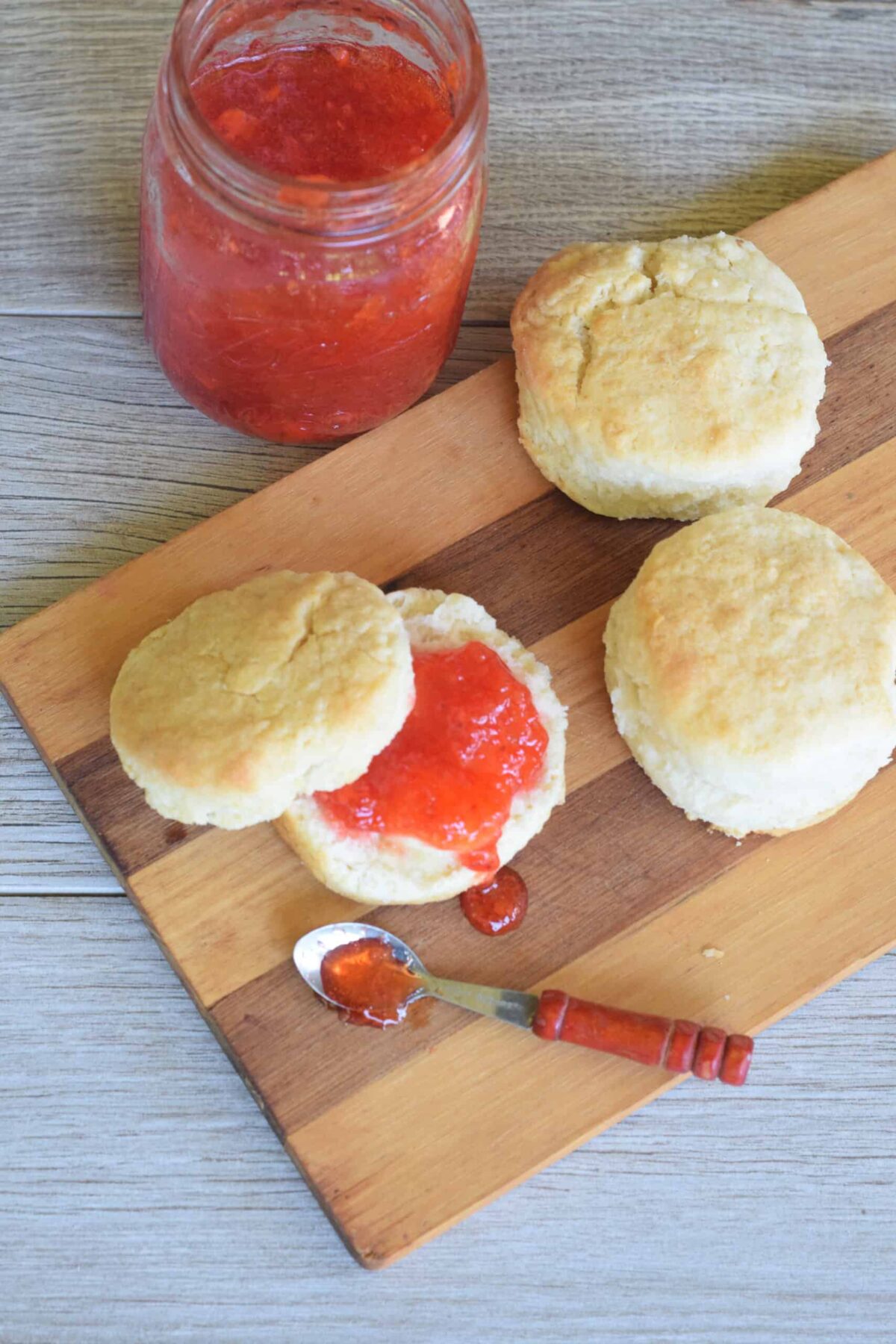 Strawberry Freezer Jam spread on 2 Ingredient Biscuits with jam spilling out of a little wooden spoon on a wooden cutting board.