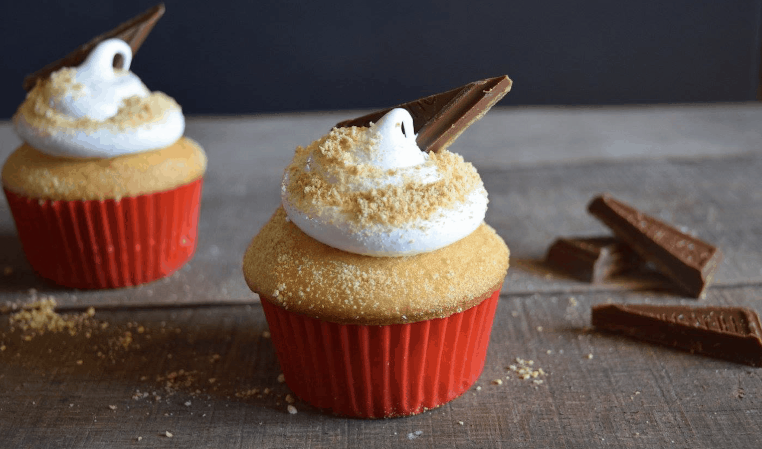 Two s'mores cupcakes with red paper lining and a triangle of chocolate bar stuck in marshmallow frosting.