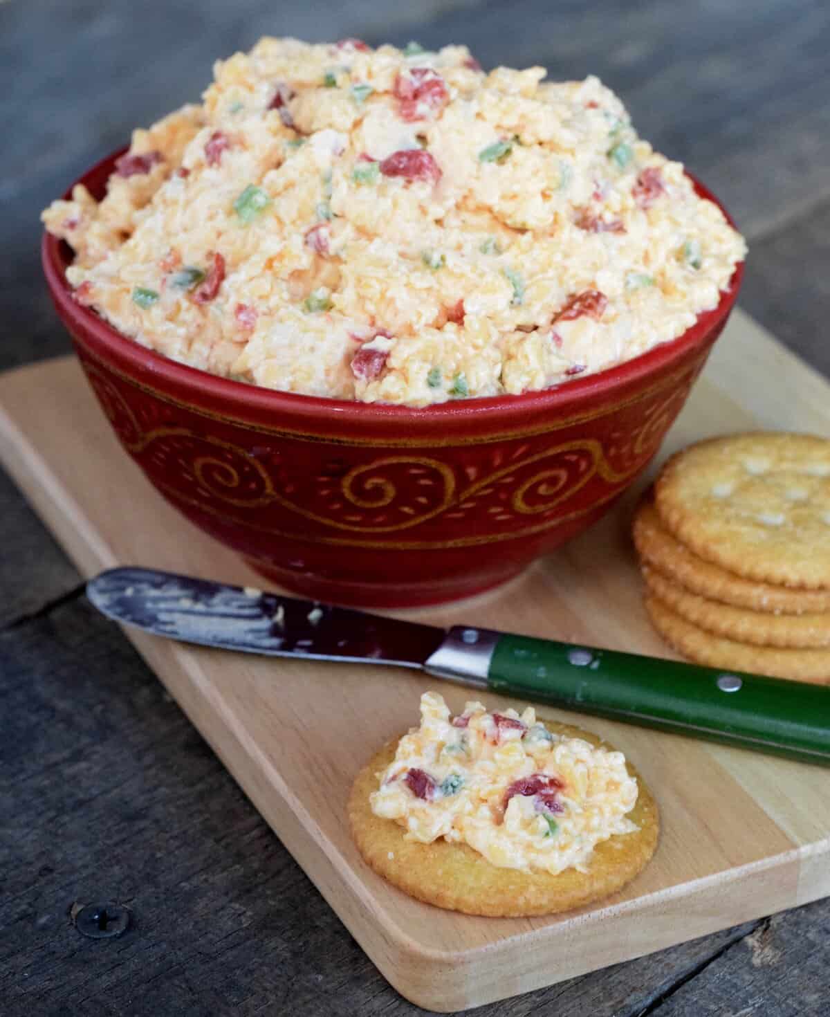 Red bowl of white pimento cheese with flecks of red and green on a wooden cutting board