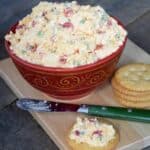 white pimento cheese spread in a red bowl on a wooden cutting board with spread on a cracker and green spreader beside it