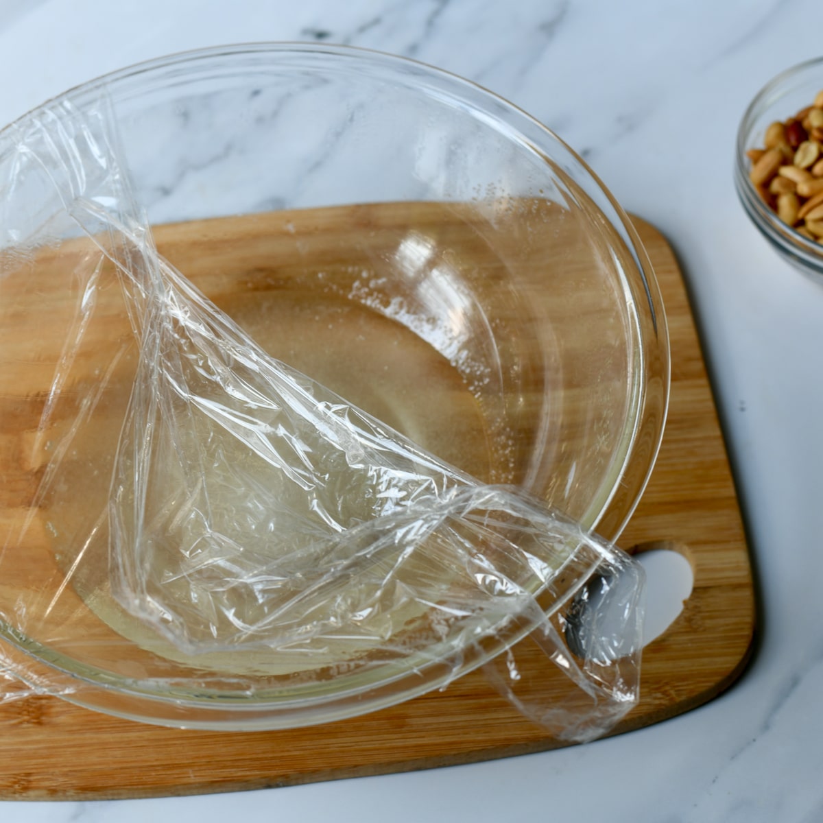 Plastic wrap halfway covering a glass bowl on top of a wooden cutting board.