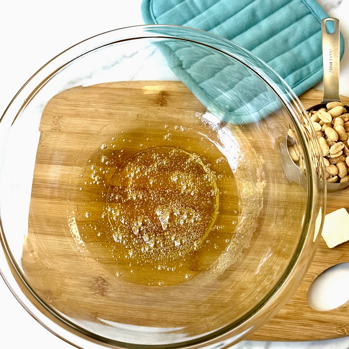 Glass bowl with golden color sugar mixture in bottom for peanut brittle on wooden cutting board.