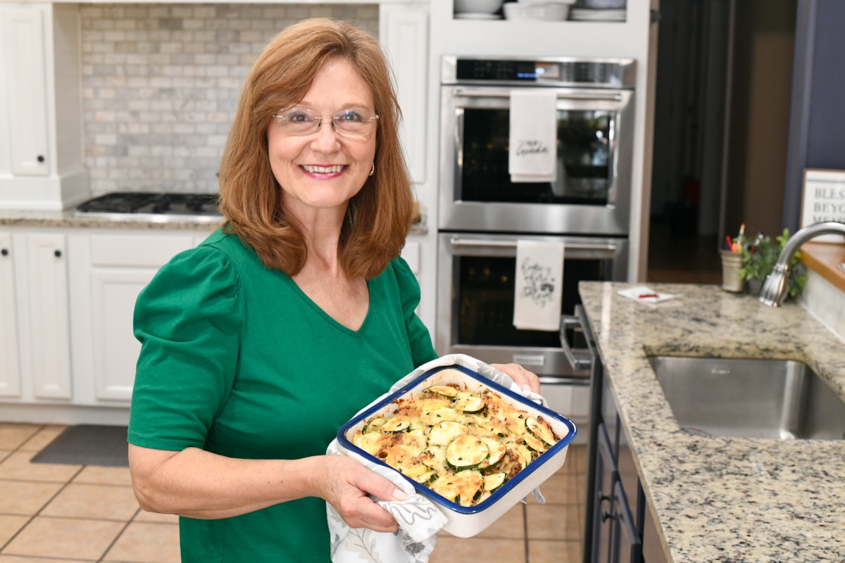 Woman holding zucchini sausage casserole with towel standing in a kitchen.