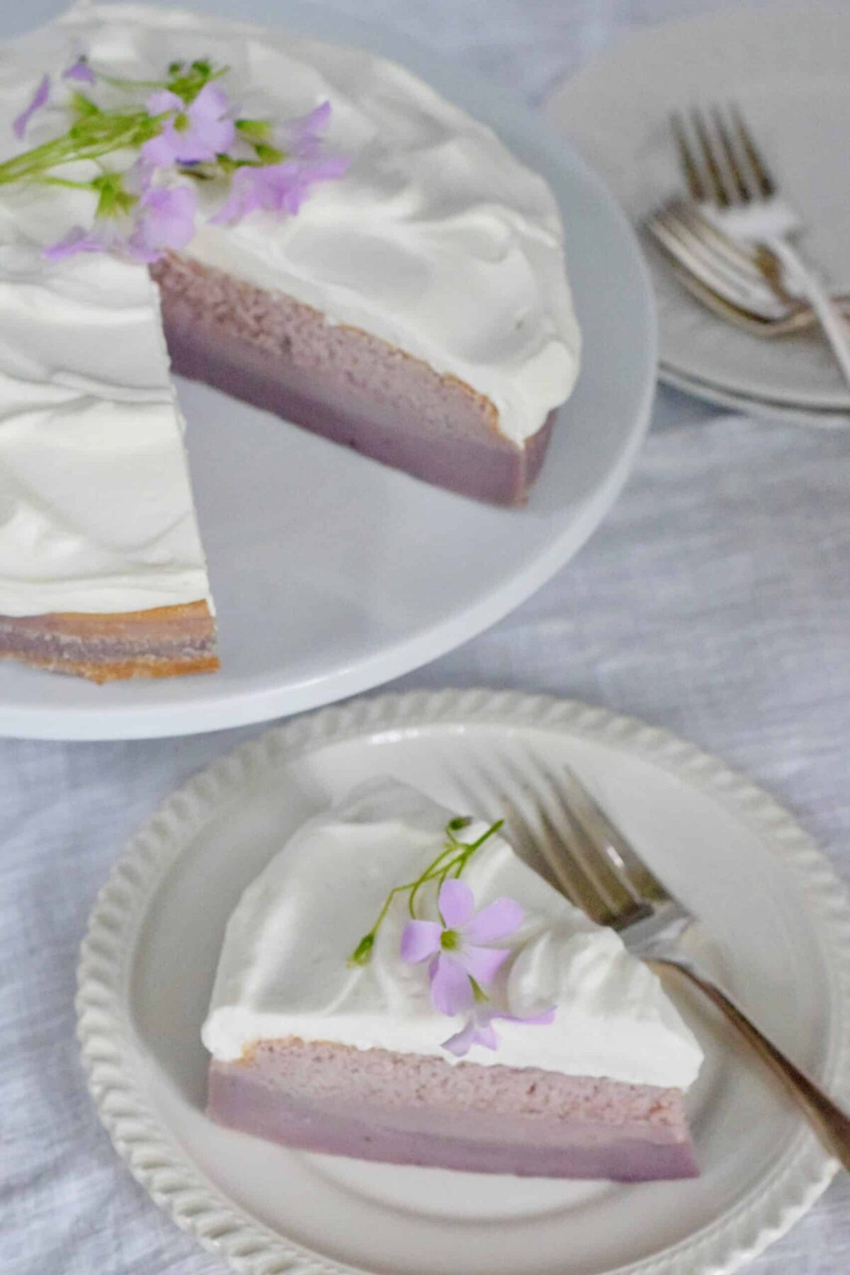 Purple Sweet potato Magic Cake with a slice cut and placed on a white plate with forks on a white tablecloth. Garnished with purple oxalis 