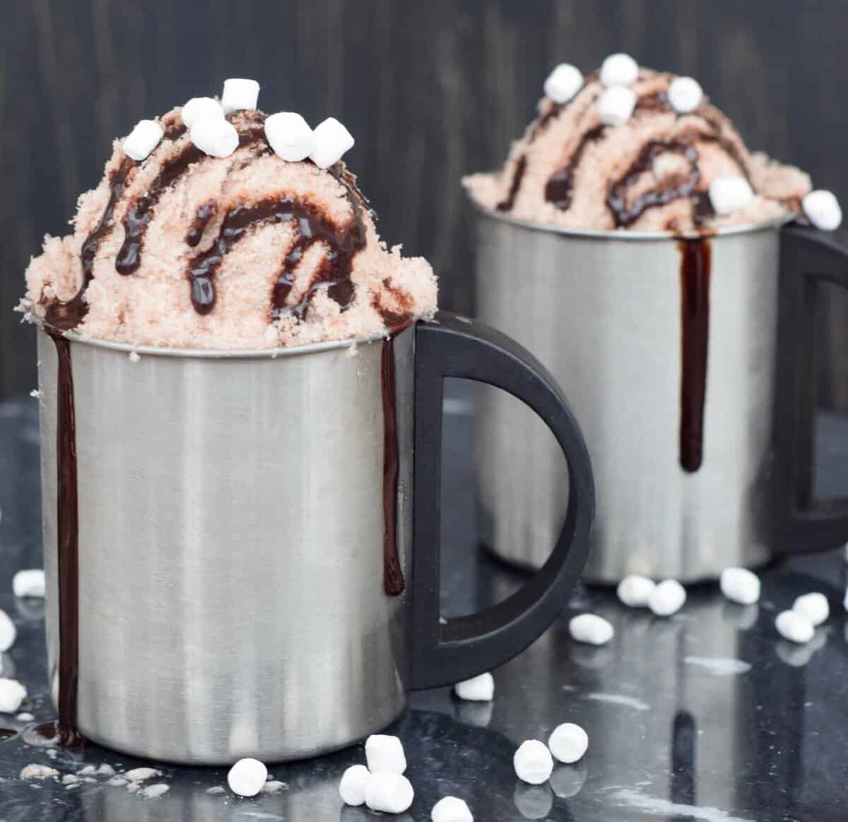 Two silver mugs of chocolate snow cream with chocolate drizzle coming down the sides and marshmallows on the black surface.