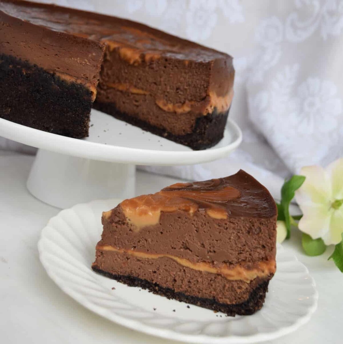 slice of chocolate cheesecake on a white plate with the rest of the cake on a cake stand