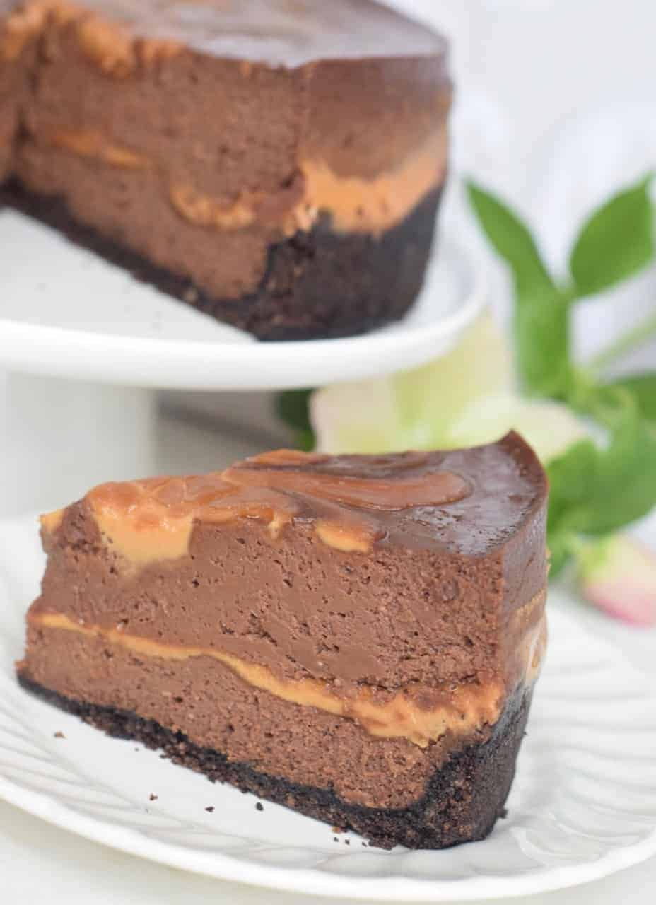 Instant Pot Choc PB Swirl Cheesecake slice on a plate with whole cheesecake on a white cake plate