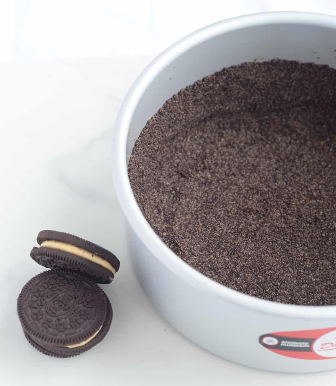 cake pan with crushed oreo crust in the bottom; two peanut butter filled sandwich cookies on the white surface