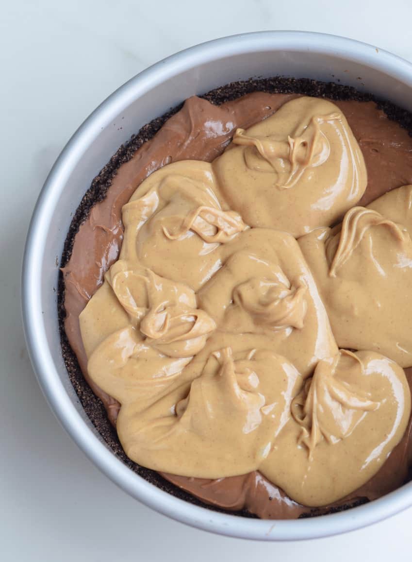 Dollopped melted peanut butter on chocolate cheesecake batter in a metal cake pan.