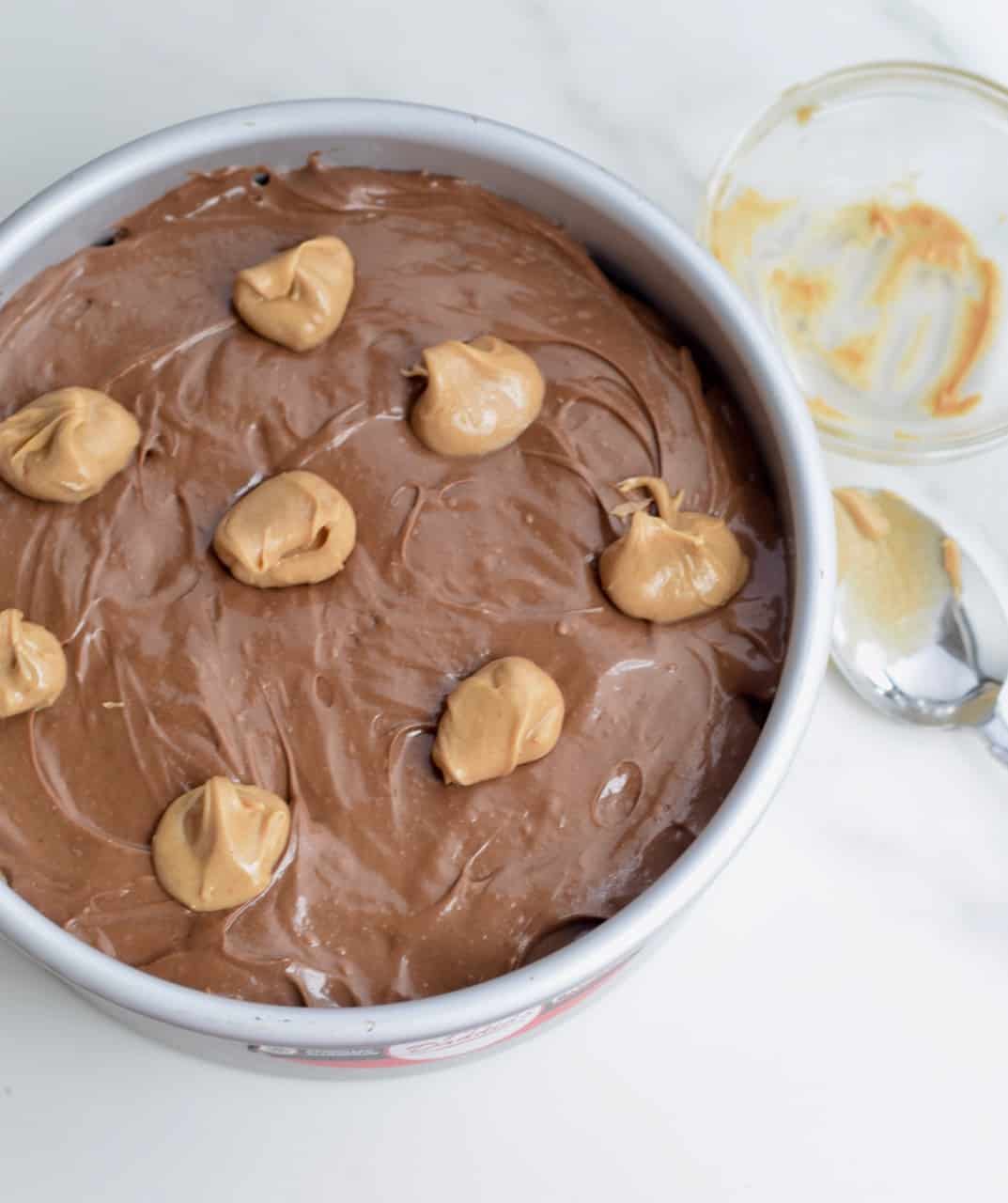 Tiny dollops of peanut butter dolloped on top of chocolate cheesecake batter in a cake pan; spoon with peanut butter on it on surface