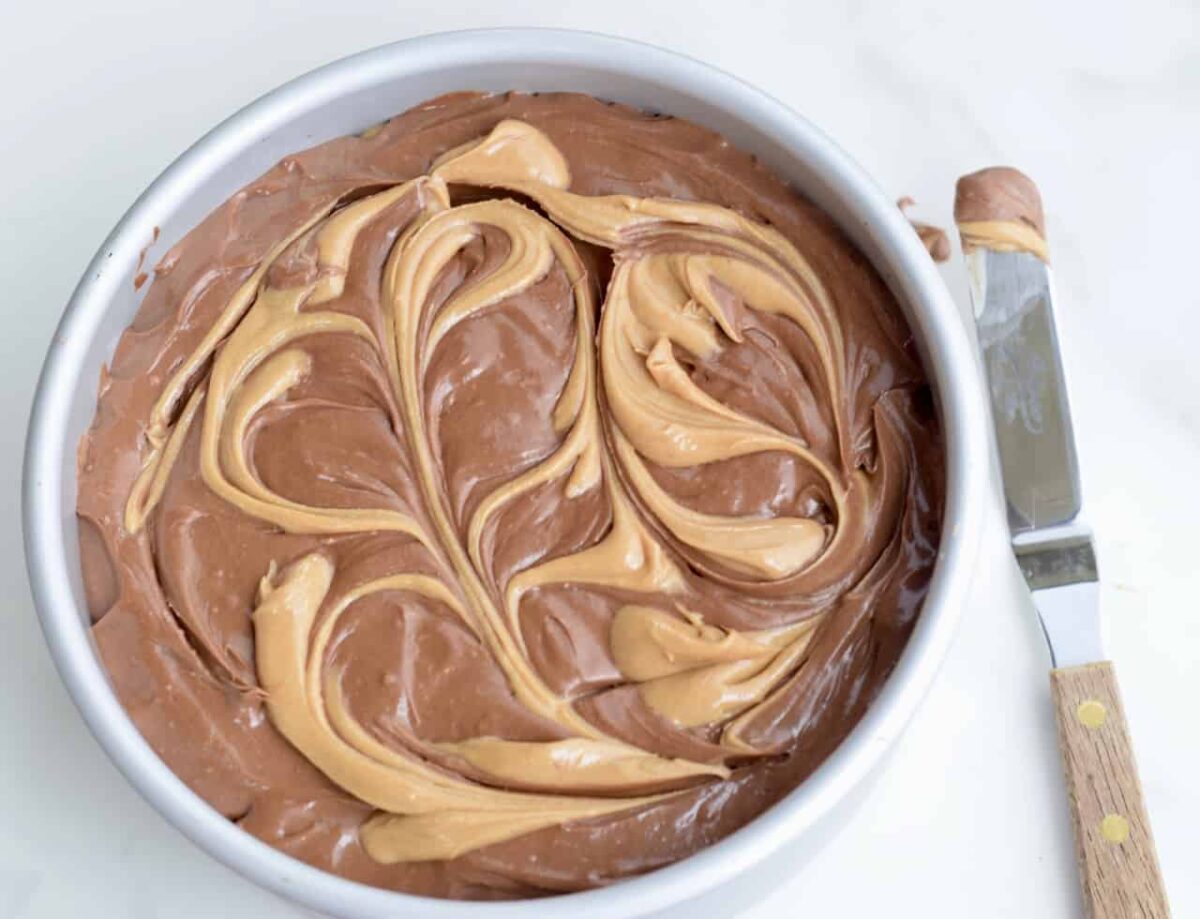 Peanut butter swirled through the top of chocolate cheesecake batter in a cake pan; spreader on surface with peanut butter on the tip.