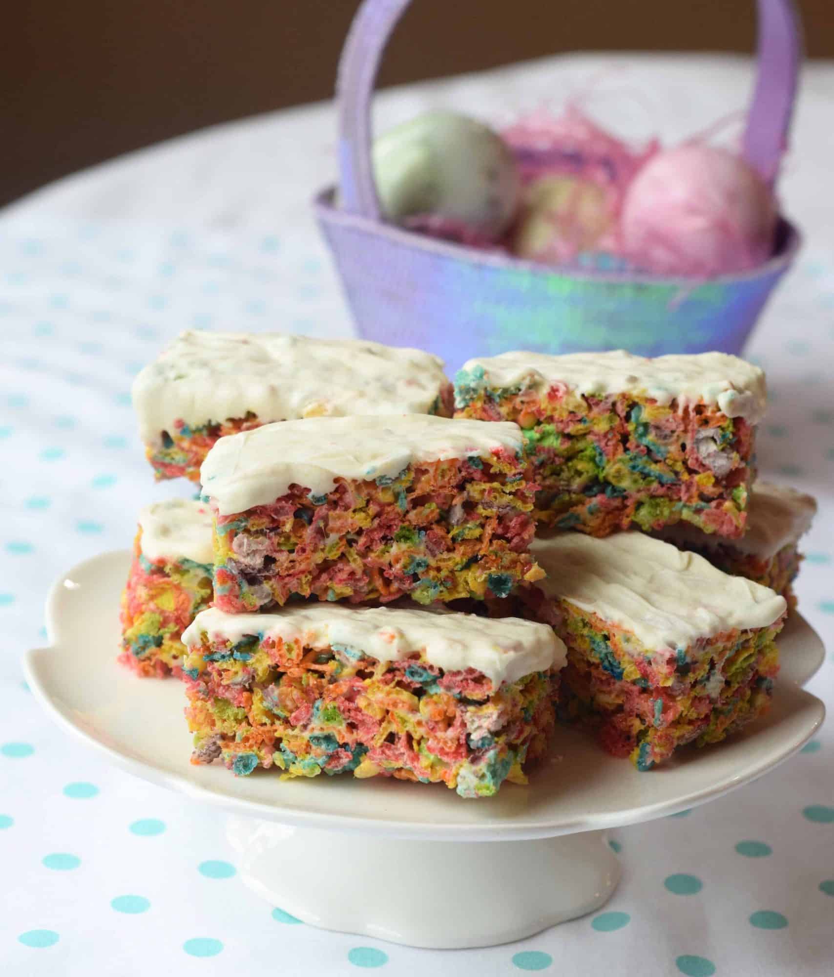 fruity rice krispy treats with white chocolate on top on a white cake stand. easter eggs in background