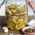 Old mason jar of pickles and onions with garlic and crushed red pepper on wooden surface