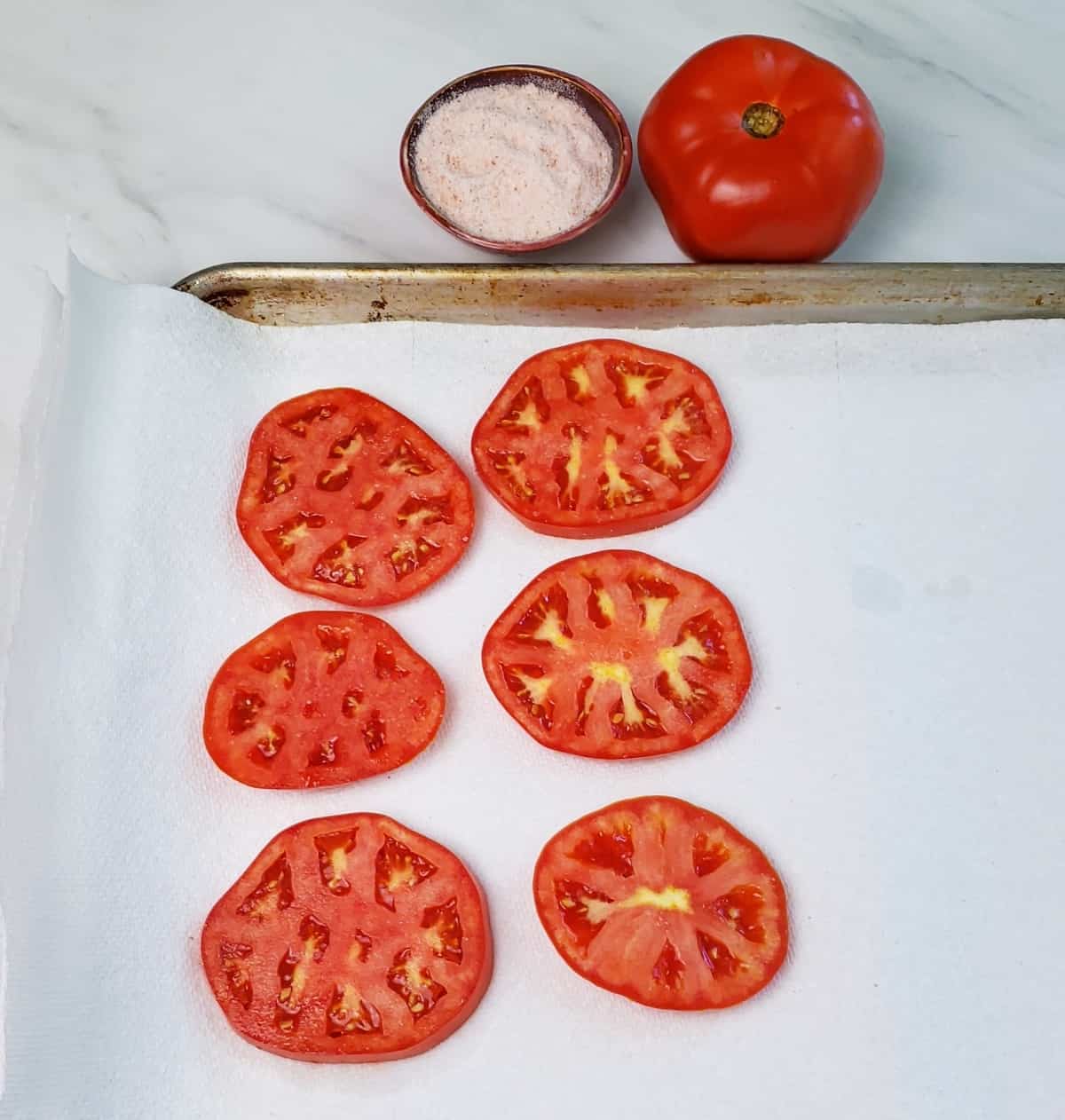 sliced tomatoes on paper towels on a baking pan. A tiny dish of pink salt and a tomato is next to pan