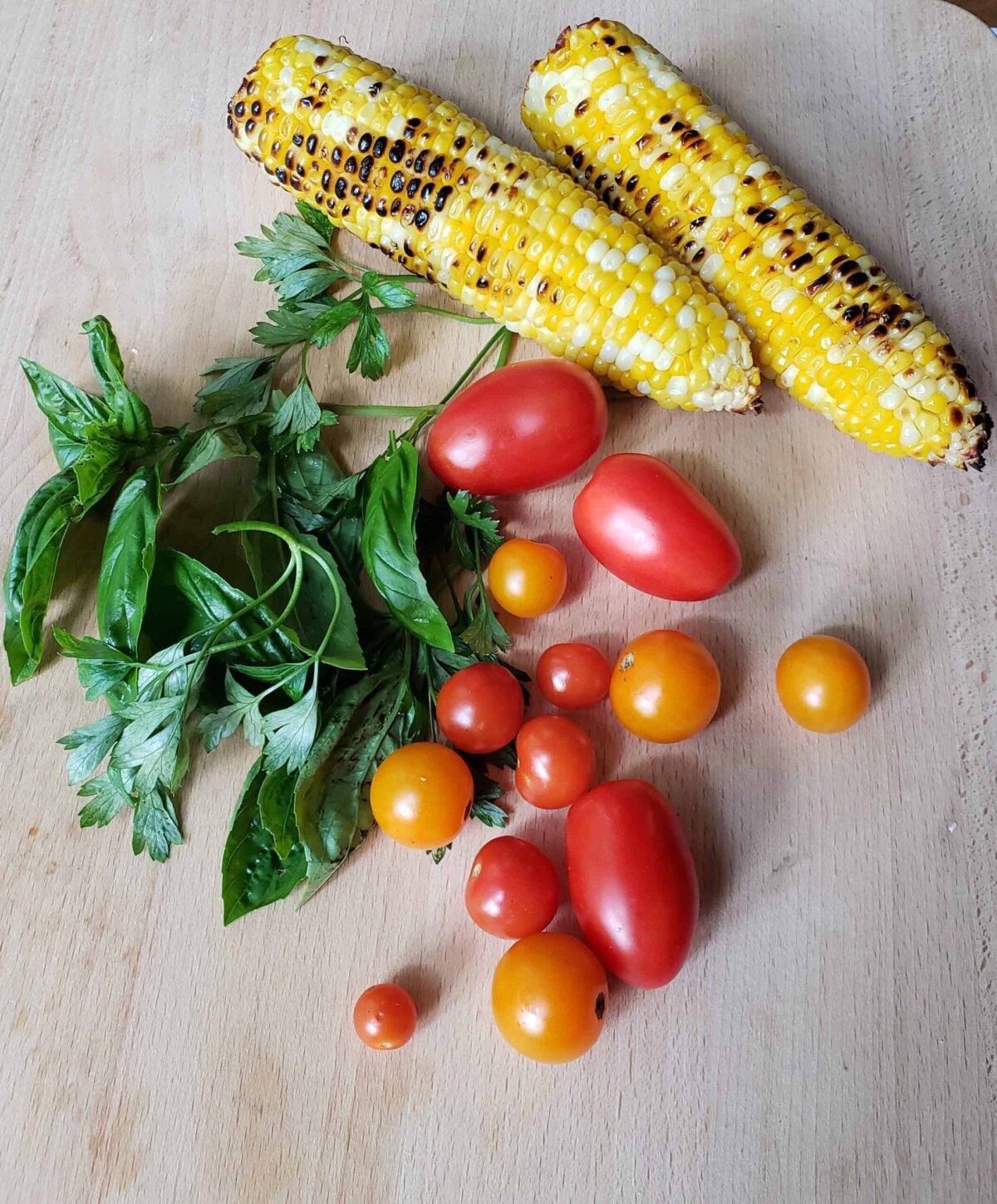 Roasted corn, red and yellow grape tomatoes, basil and parsley on wooden surface