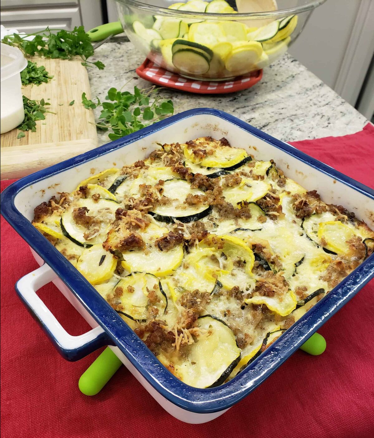 Sausage squash casserole on green silicone trivet on red surface with fresh herbs on cutting board in background