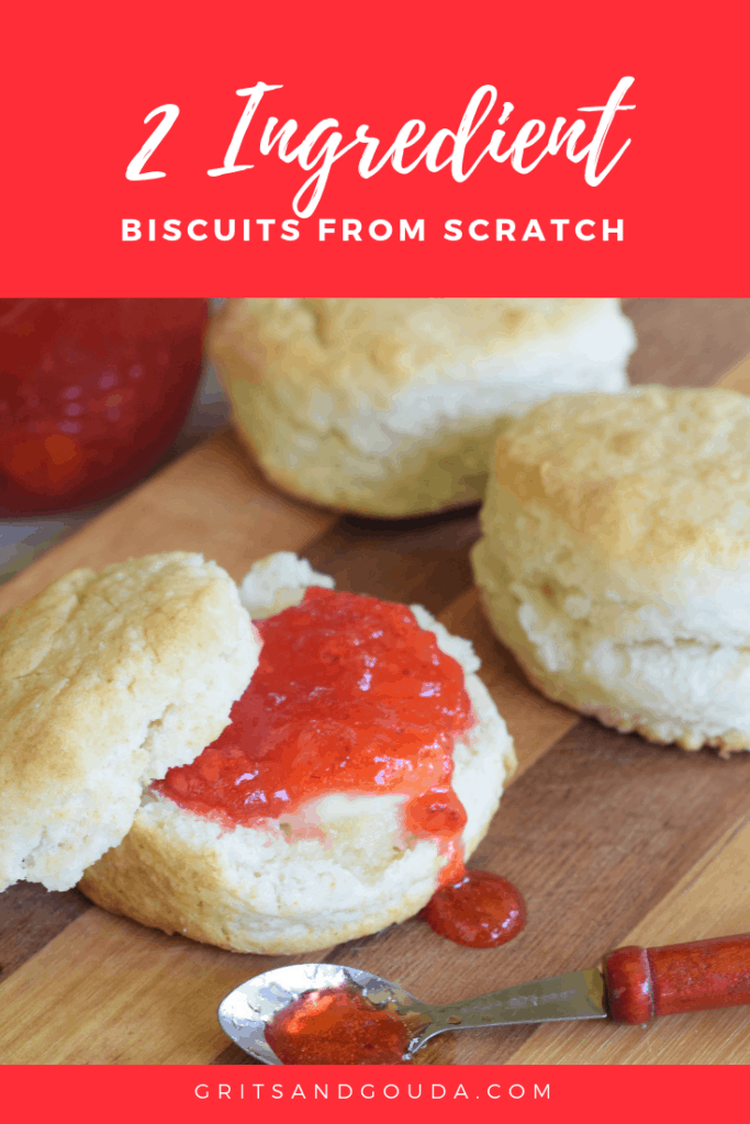 3 biscuits on a wooden cutting board spread with butter and strawberry jam. Tiny spoonful of jam on a spoon spilling out.