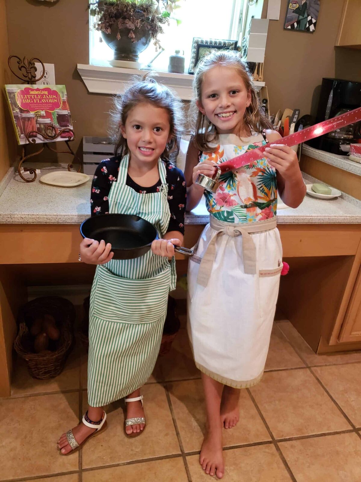 Two young girls holding a cast iron skillet and ruler and biscuit cutter wearing aprons