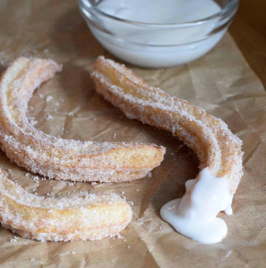 Churros with vanilla icing dipping sauce on brown parchment paper and bowl of dipping sauce