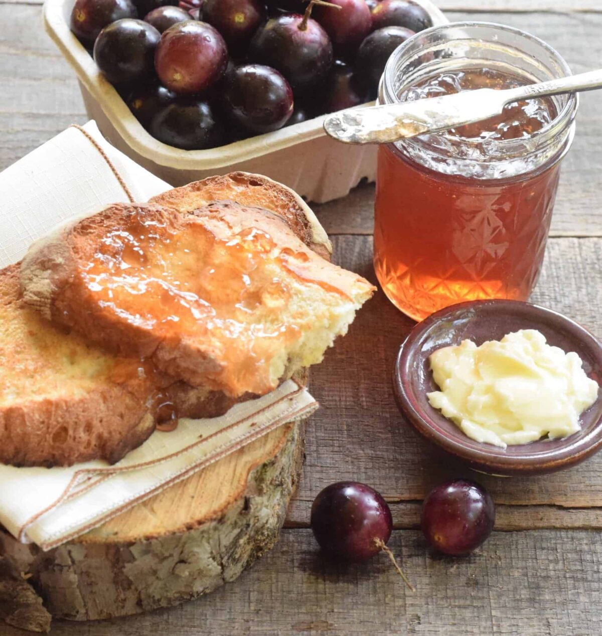 pint of muscadine grapes, jar of scuppernong jelly with knife resting on it, buttered slices of bread with jelly on a napkin on a slice of wood, butter in a ceramic plate