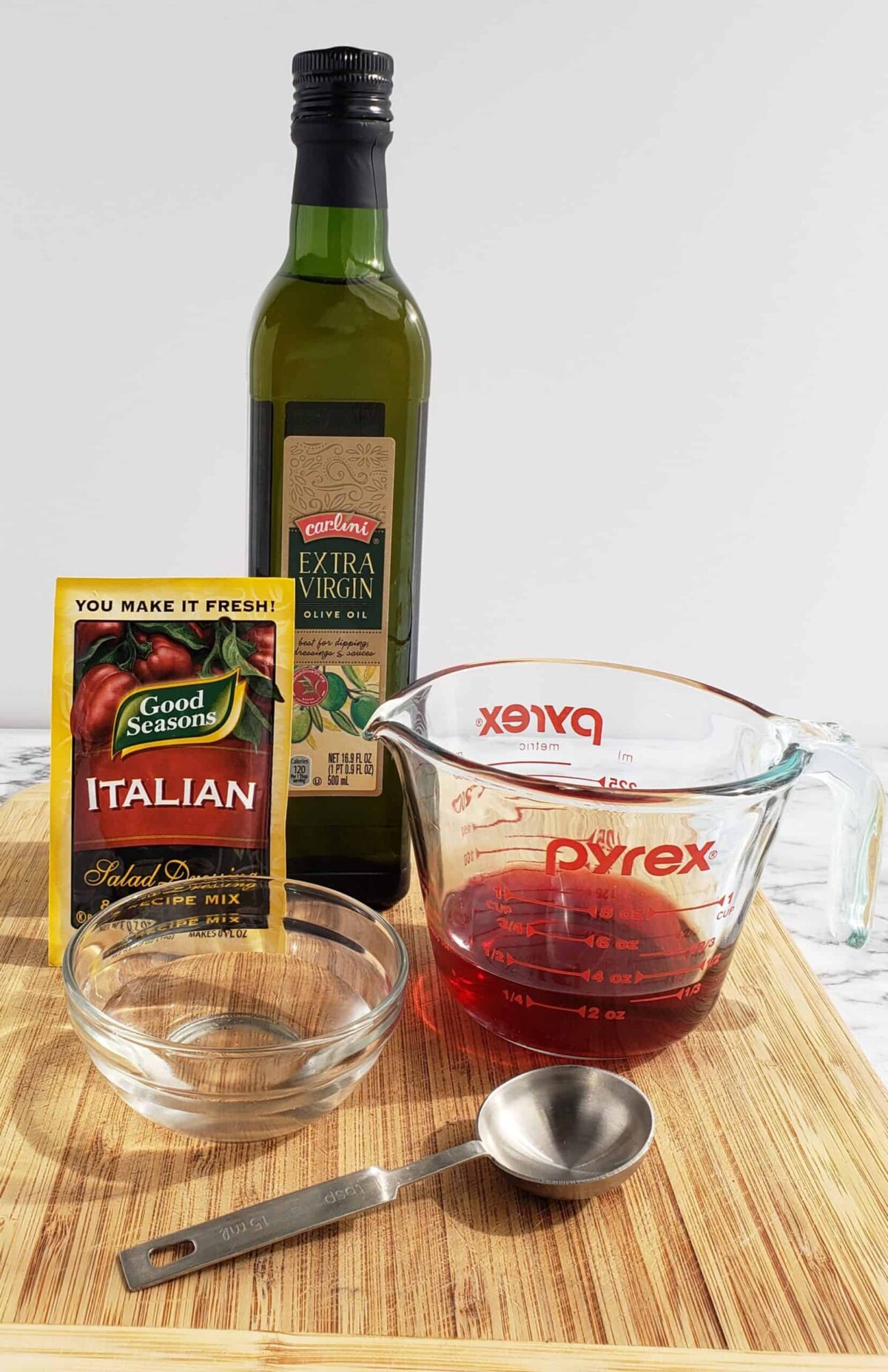 bottle of olive oil, measuring cup with blackberry vinegar, metal tablespoon, little glass bowl, package of dry Italian Salad dressing