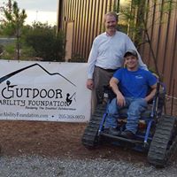 Scott Phillips and Grayson Phillips next to the Outdoor Ability Foundation banner. Grayson is in an Action Trackchair