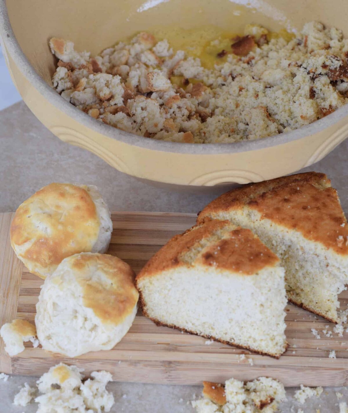 cornbread slices and 2 biscuits on wooden cutting board with bread crumbled in bowl
