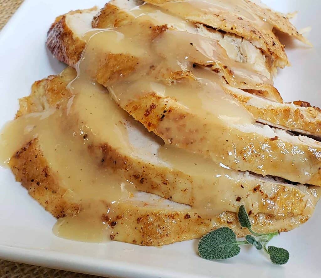 Slices of turkey breast with skin on overlapping each other on a white plate and garnished with fresh sage and gravy poured over the turkey slices