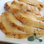 Slices of turkey breast with skin on overlapping each other on a white plate and garnished with fresh sage and gravy poured over the turkey slices
