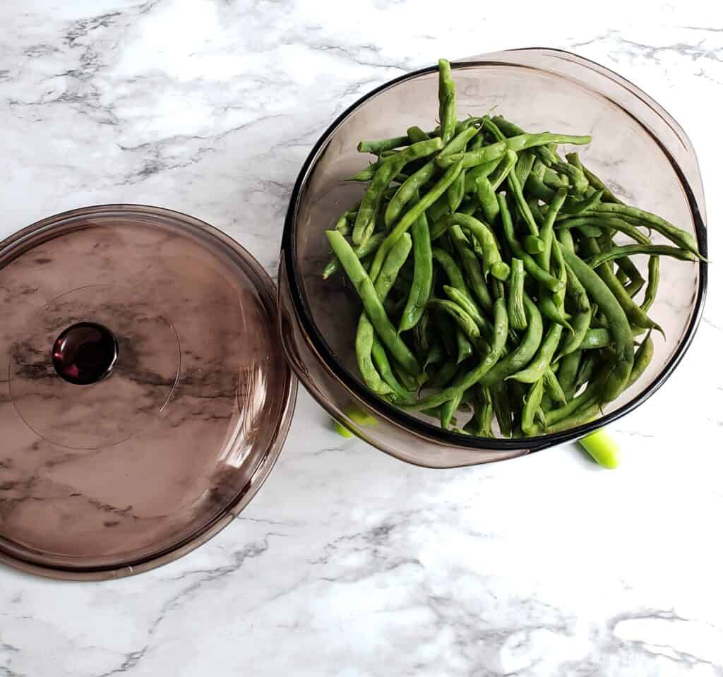 Fresh green beans steamed in a dark pyrex bowl with the lid on a marble surface.