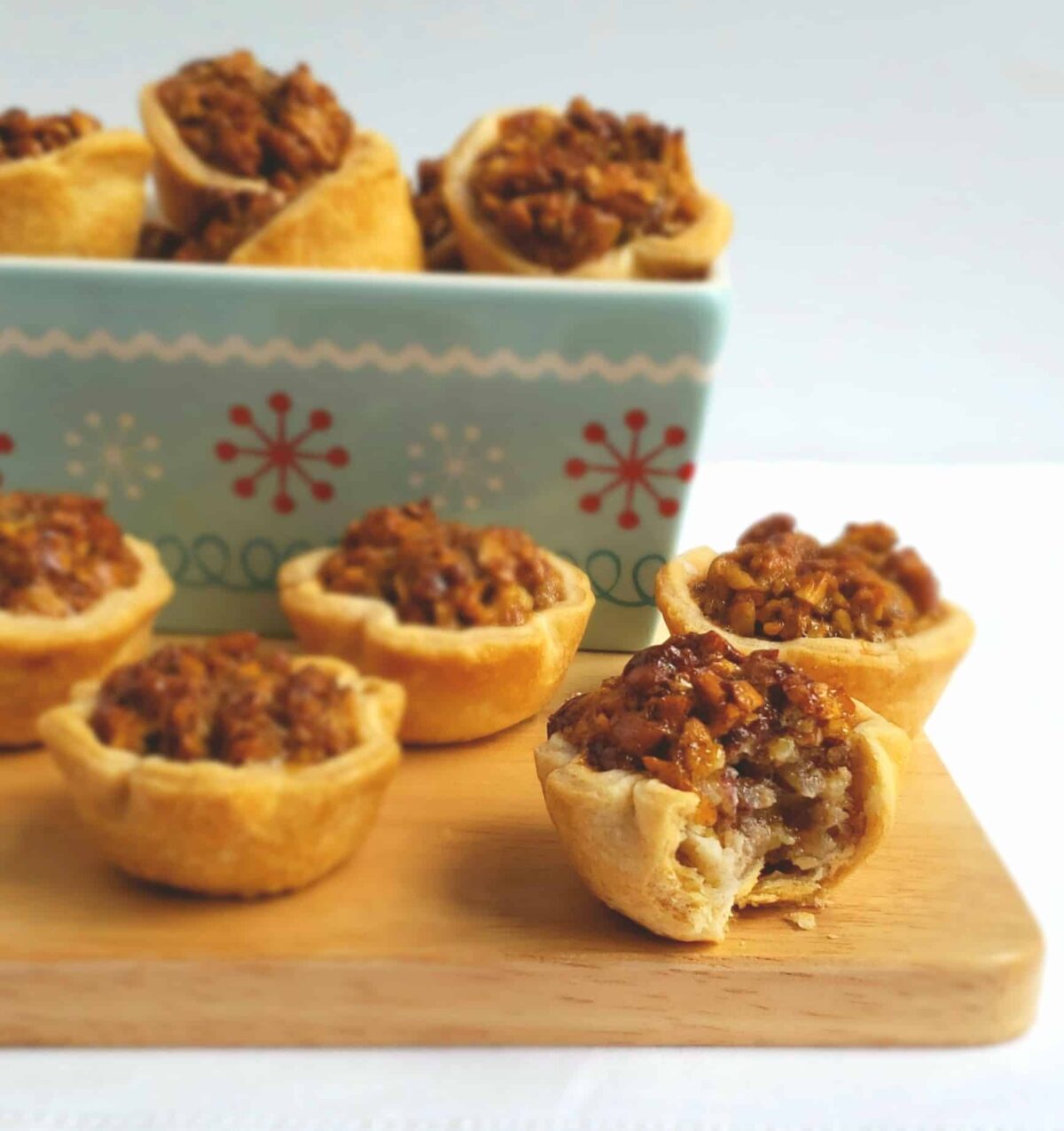 Maple Pecan Tassies are mini pecan pies. They are arranged on a wooden board and in a snowflake rectangle dish