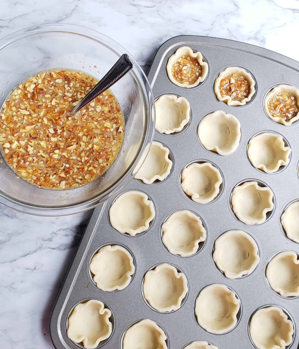 Rounds of refrigerated pie dough fitted in mini muffin pans with a bowl of Maple Pecan Pie filling next to it.