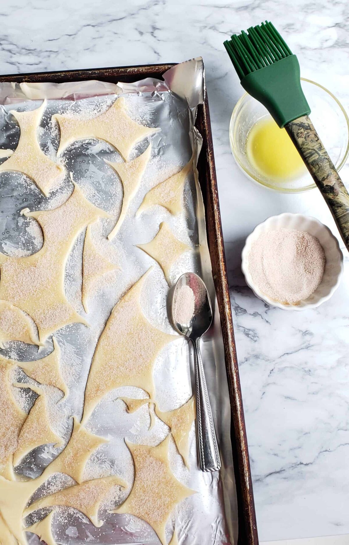 Scraps from cutting out pie crust dough on a foil-lined baking sheet with bowl of cinnamon sugar and pastry brush with bowl of melted butter