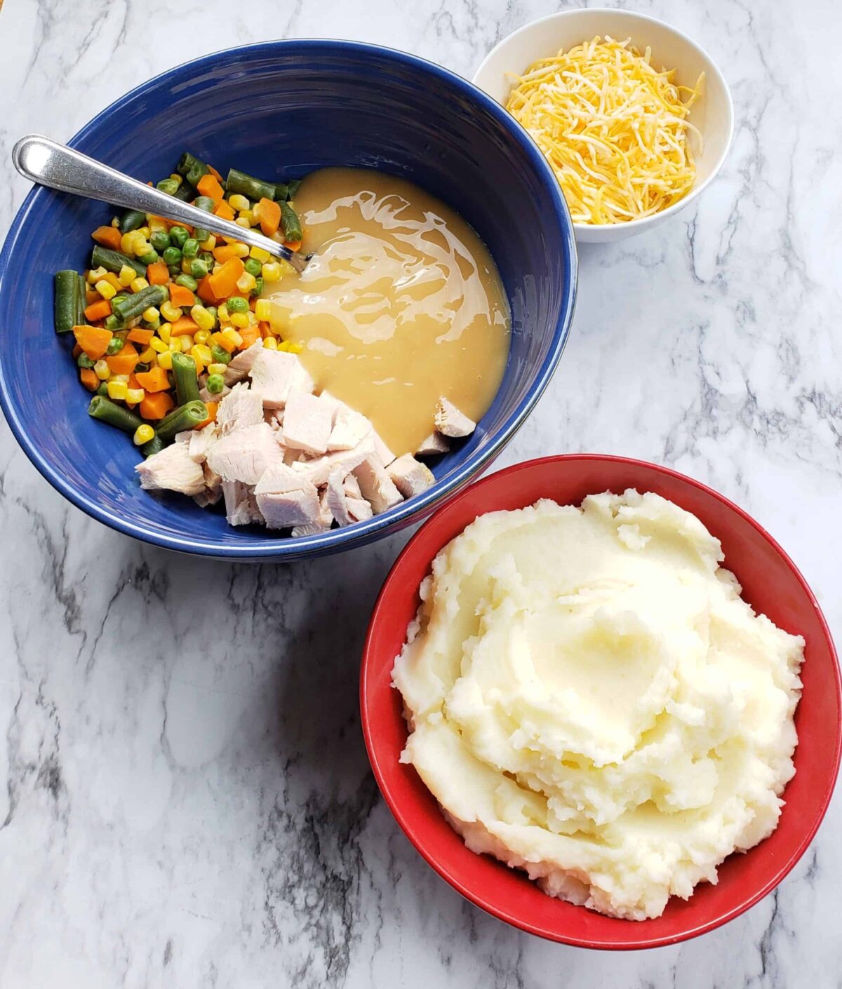 Blue bowl filled with mixed vegetables, chopped turkey, and gravy. Red bowl of mashed potatoes. small white bowl of shredded cheese.