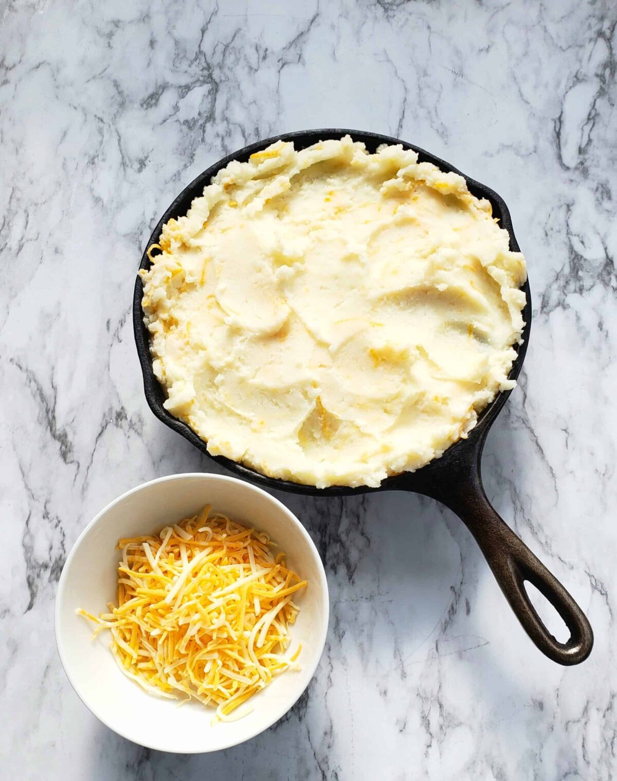 Cast iron skillet full of mashed potatoes. Small white bowl of shredded cheese