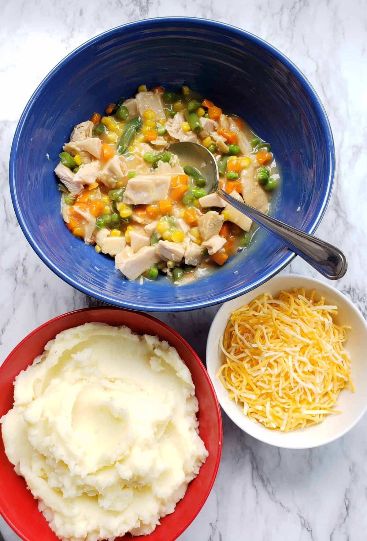 Chopped turkey, vegetables, and gravy in a blue bowl. Mashed potatoes in a red bowl. Shredded cheese in a small white bowl.
