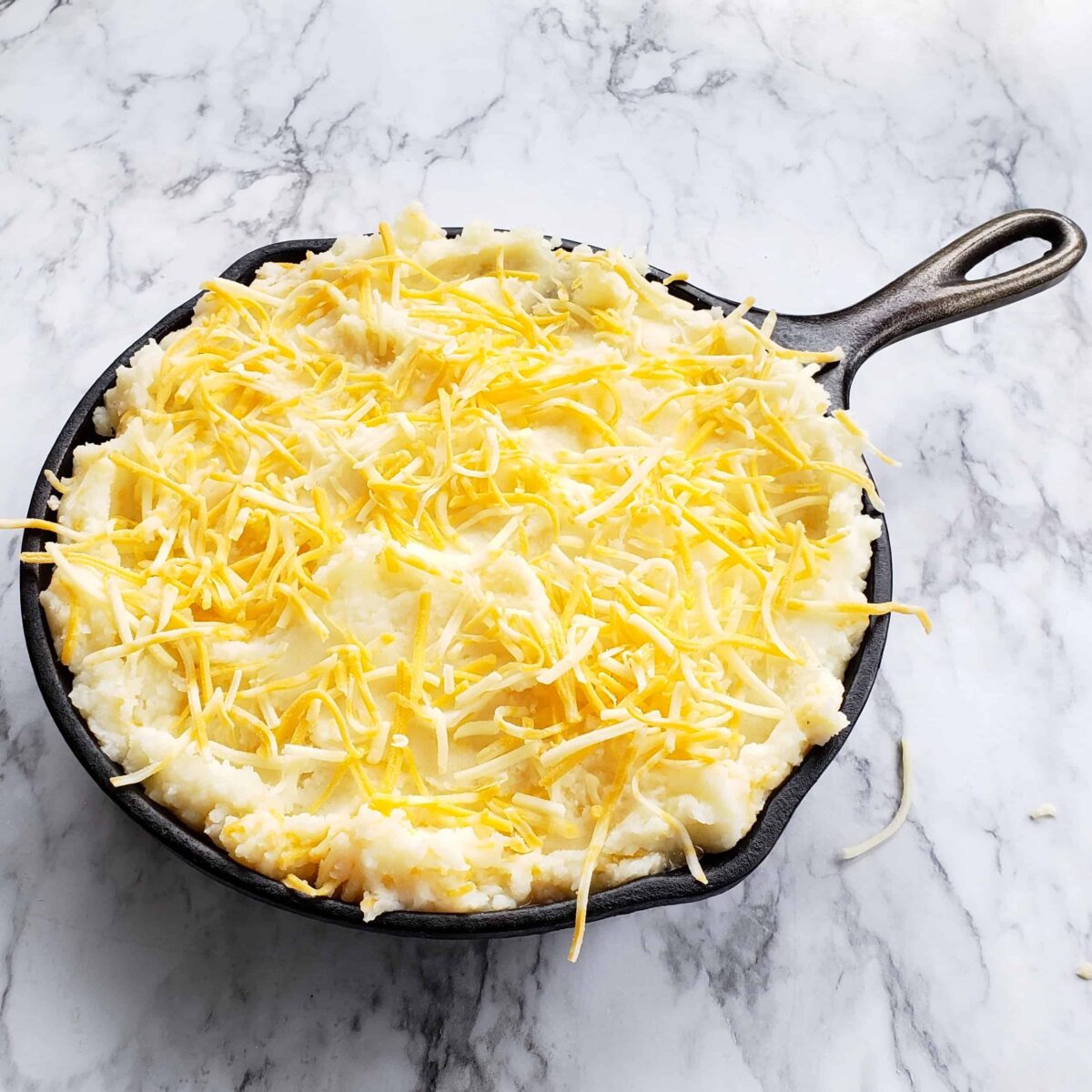 Cast iron skillet full of mashed potatoes with Colby Jack cheese sprinkled on top on marble surface