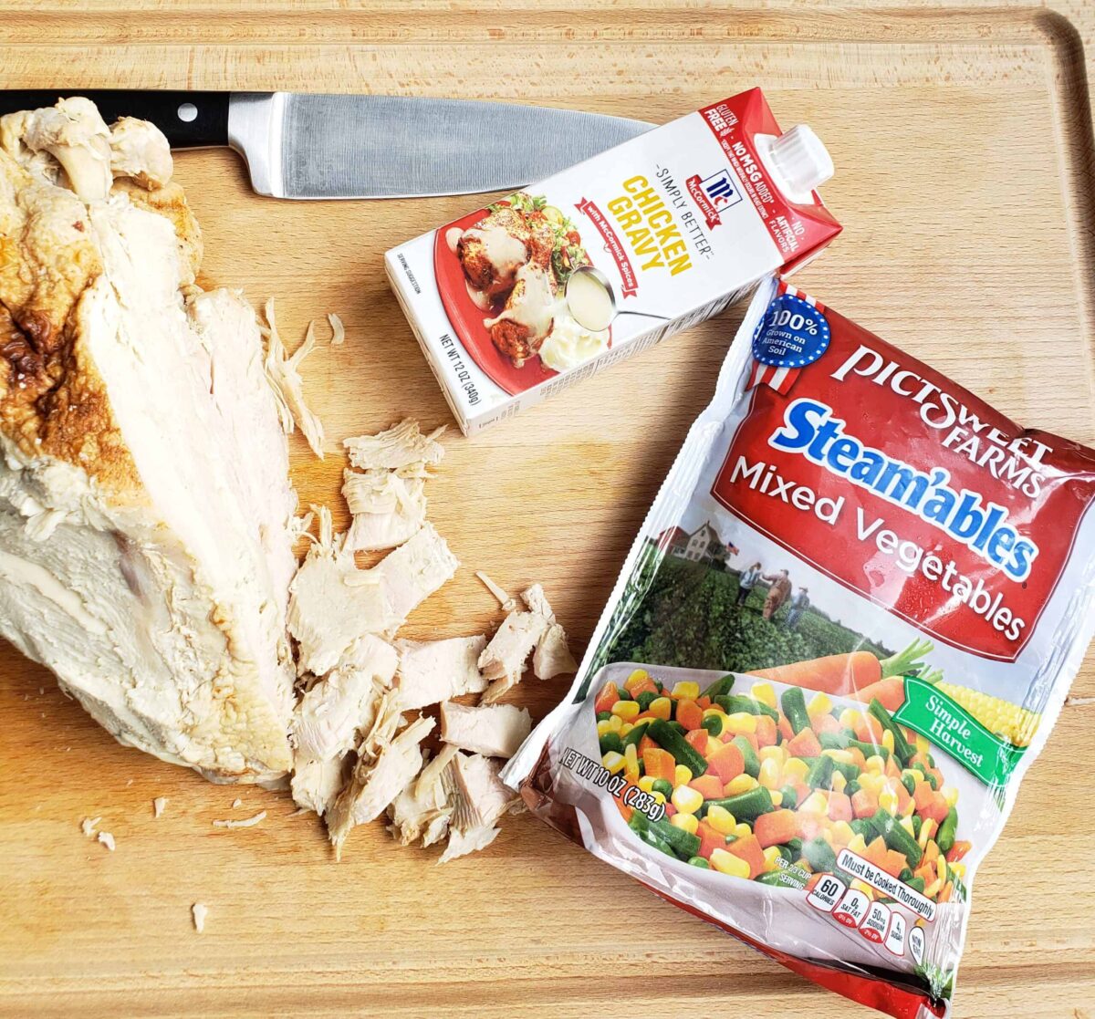 Turkey breast with some cut off the breast, a chefs knife on a cutting board, McCormicks chicken gravy and PictSweet mixed vegetables in package