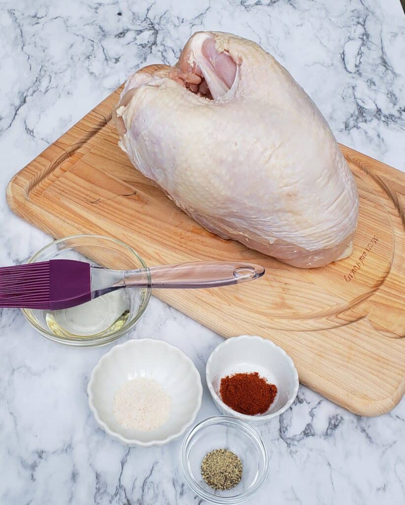Uncooked turkey breast with dishes of spices and a tiny bowl of oil with a pastry brush on a marble surface.