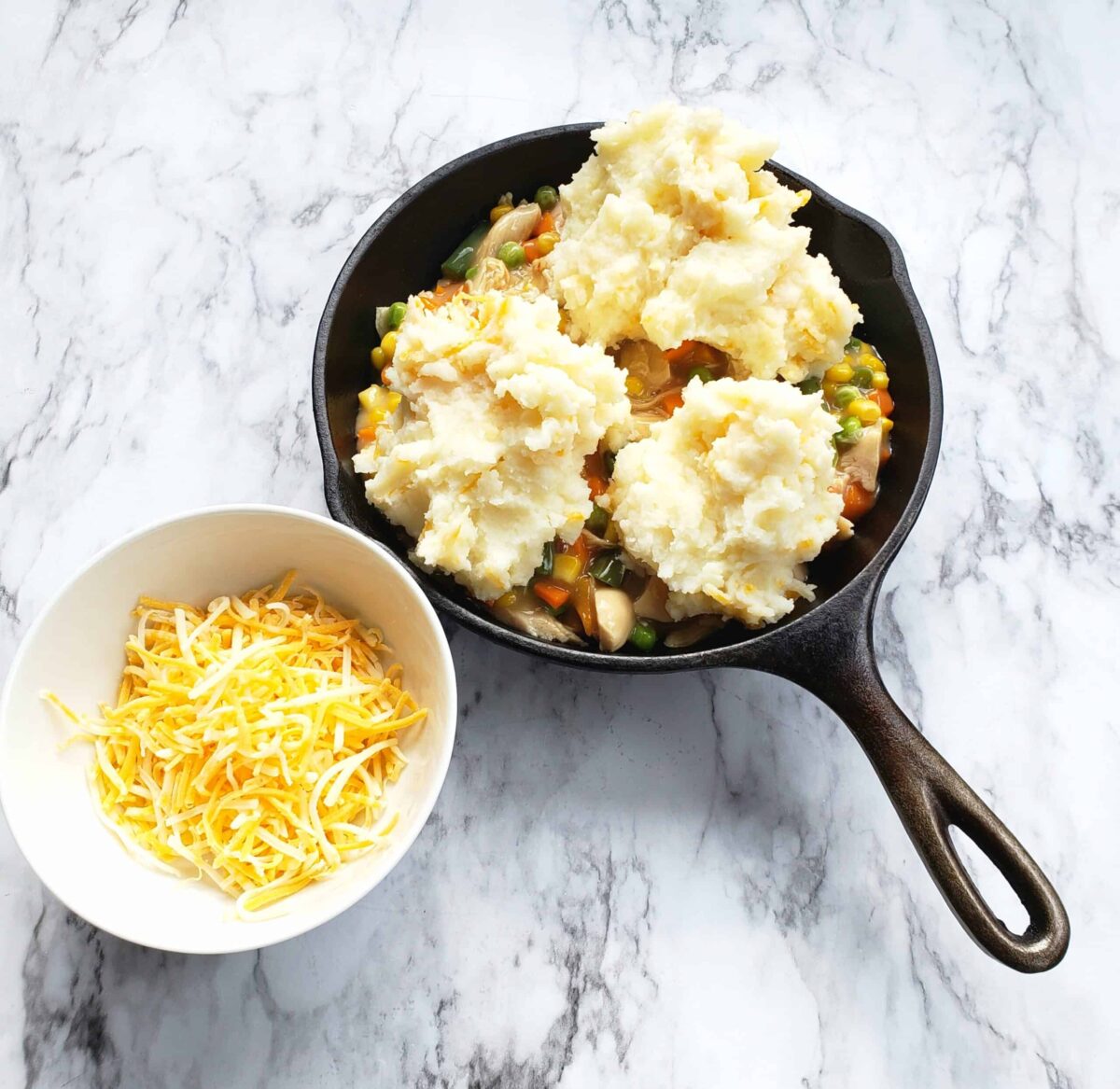 Vegetables and gravy and turkey in a cast iron skillet with mashed potatoes dolloped on top. Small white bowl of shredded cheese