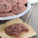 Double Chocolate Chip Cookies are soft and chewy from a secret ingredient. A bite is taken out of this one and placed on a piece of parchment. A glass of milk is close by to wash them down.