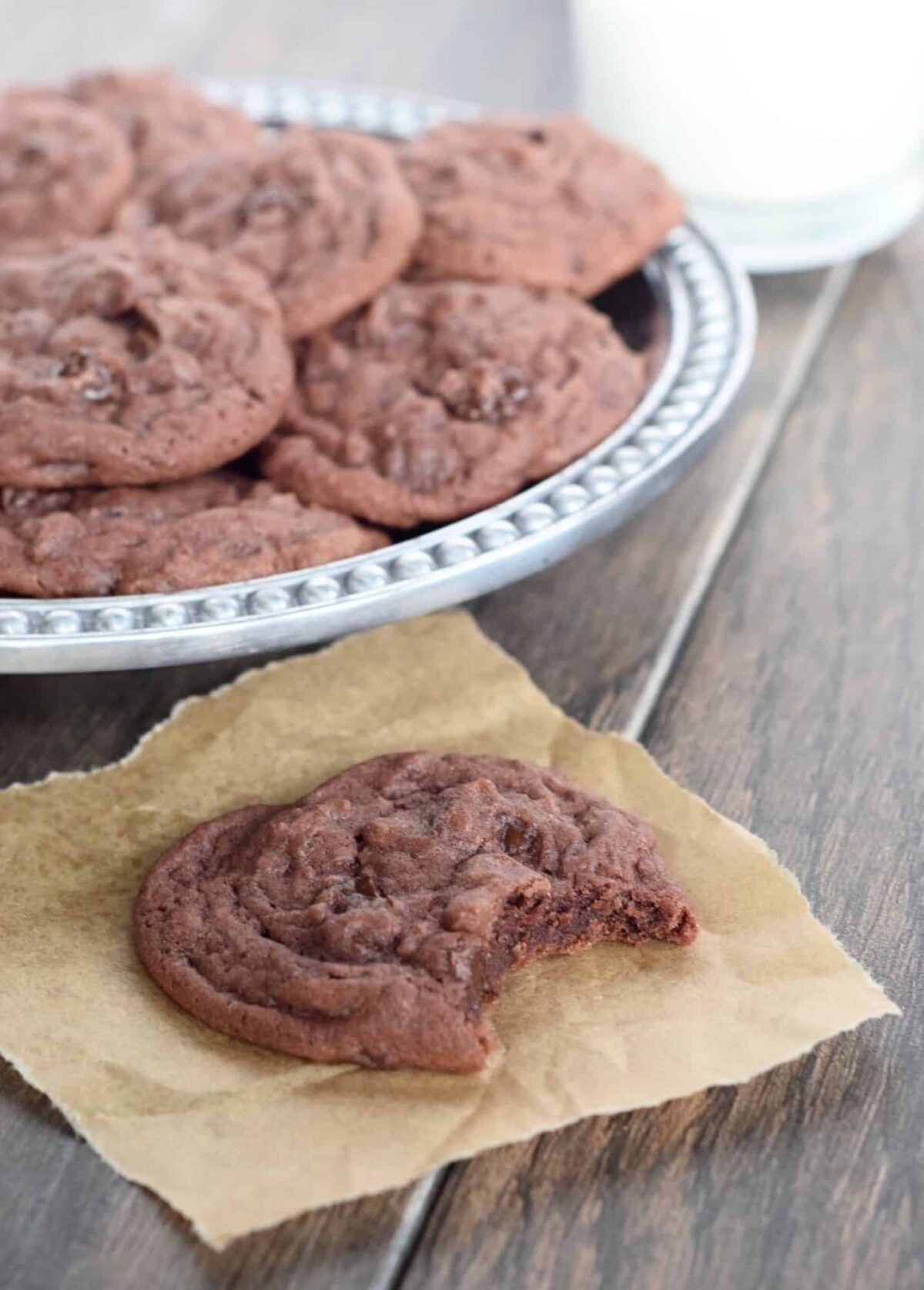 Chocolate cookies with chocolate chips with bite out of one on parchment. Plate of them behind it and glass of milk