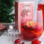 stemless wine class of sangria with pink box of tea and sliver Christmas beads on white surface