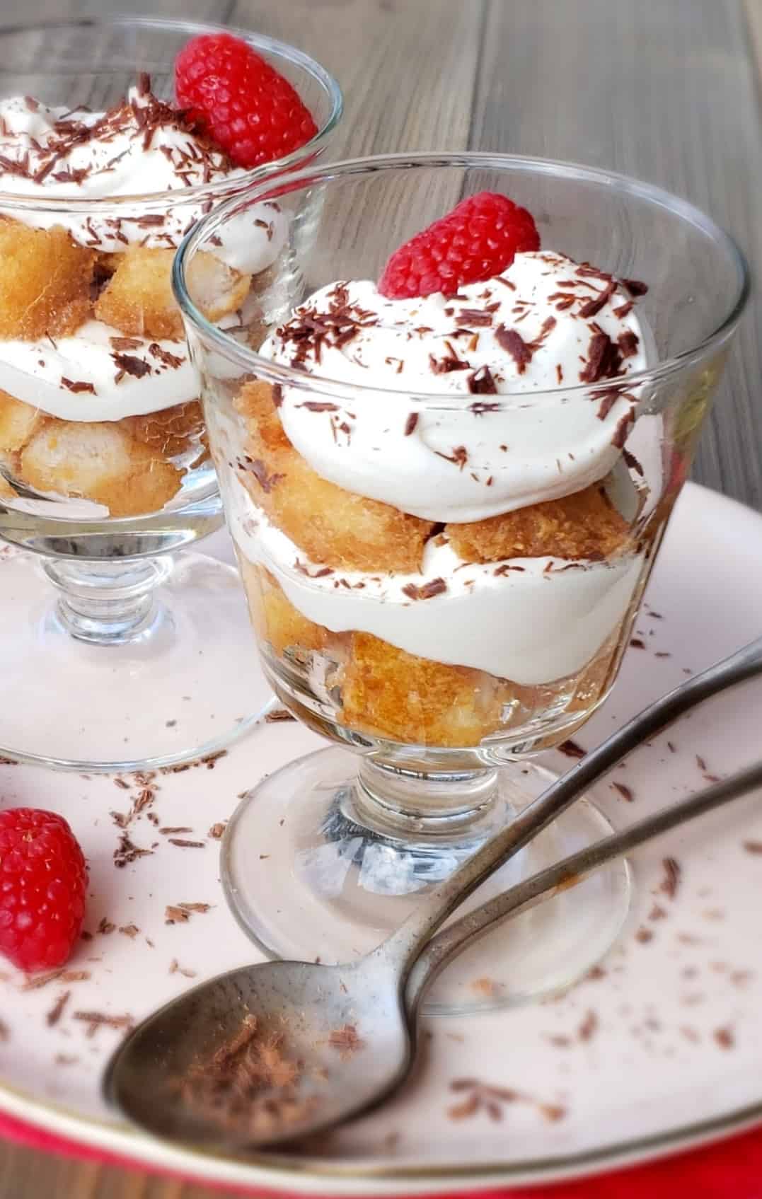 layered Tiramisu in two glass dessert dishes with raspberries and chocolate shavings on a pink plate with tea spoons