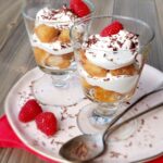 layered Tiramisu in two glass dessert dishes with raspberries and chocolate shavings on a pink plate with tea spoons