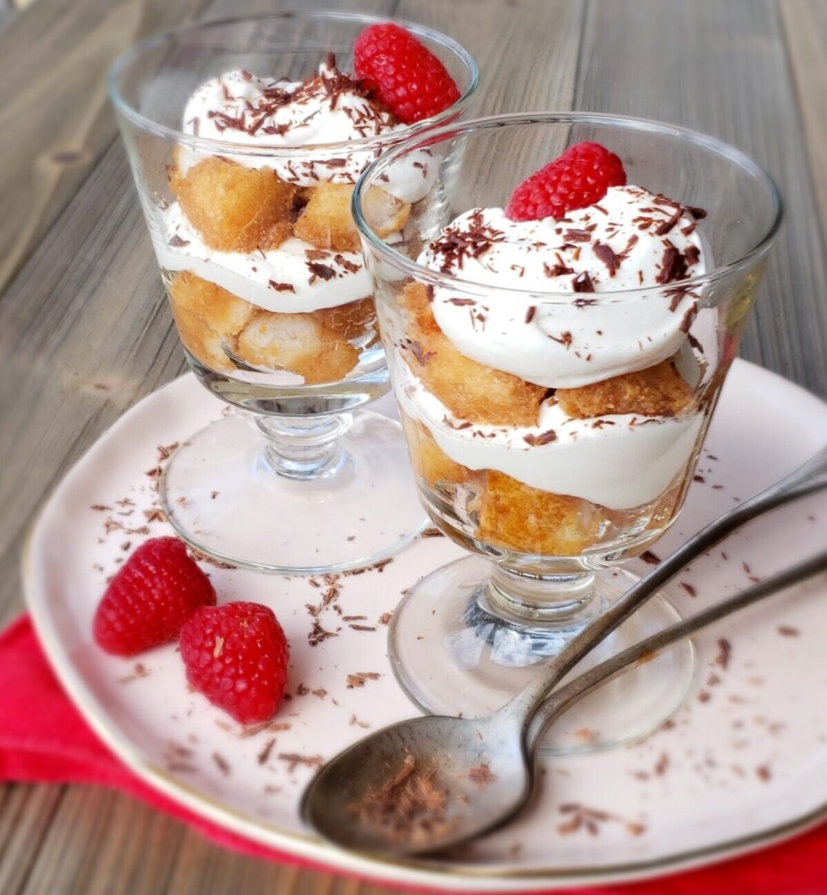 layered Tiramisu in two glass dessert dishes with raspberries and chocolate shavings on a pink plate with tea spoons