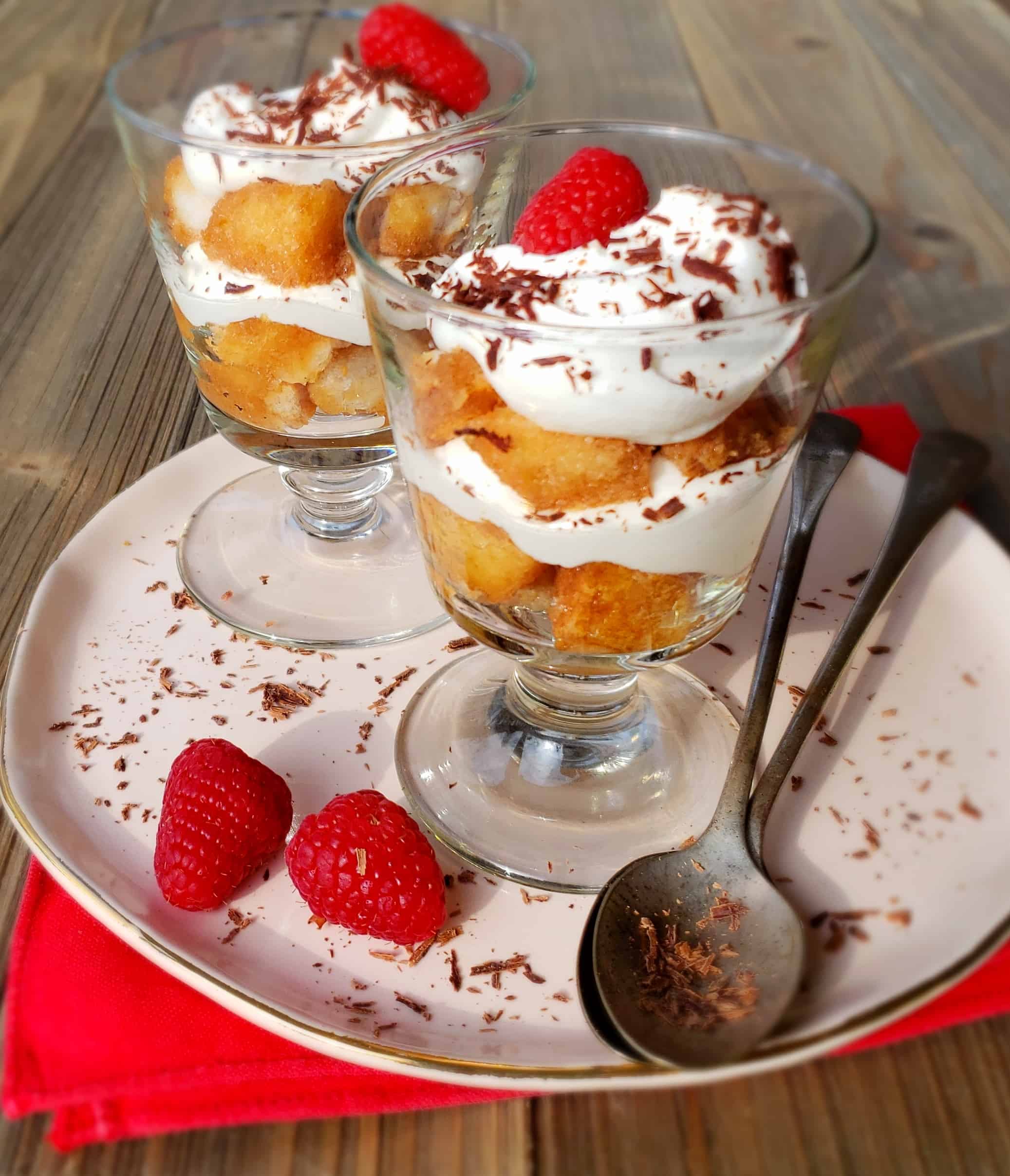 Two parfait glasses layered with tea soaked cake, whipped cream, and topped with raspberries and grated chocolate